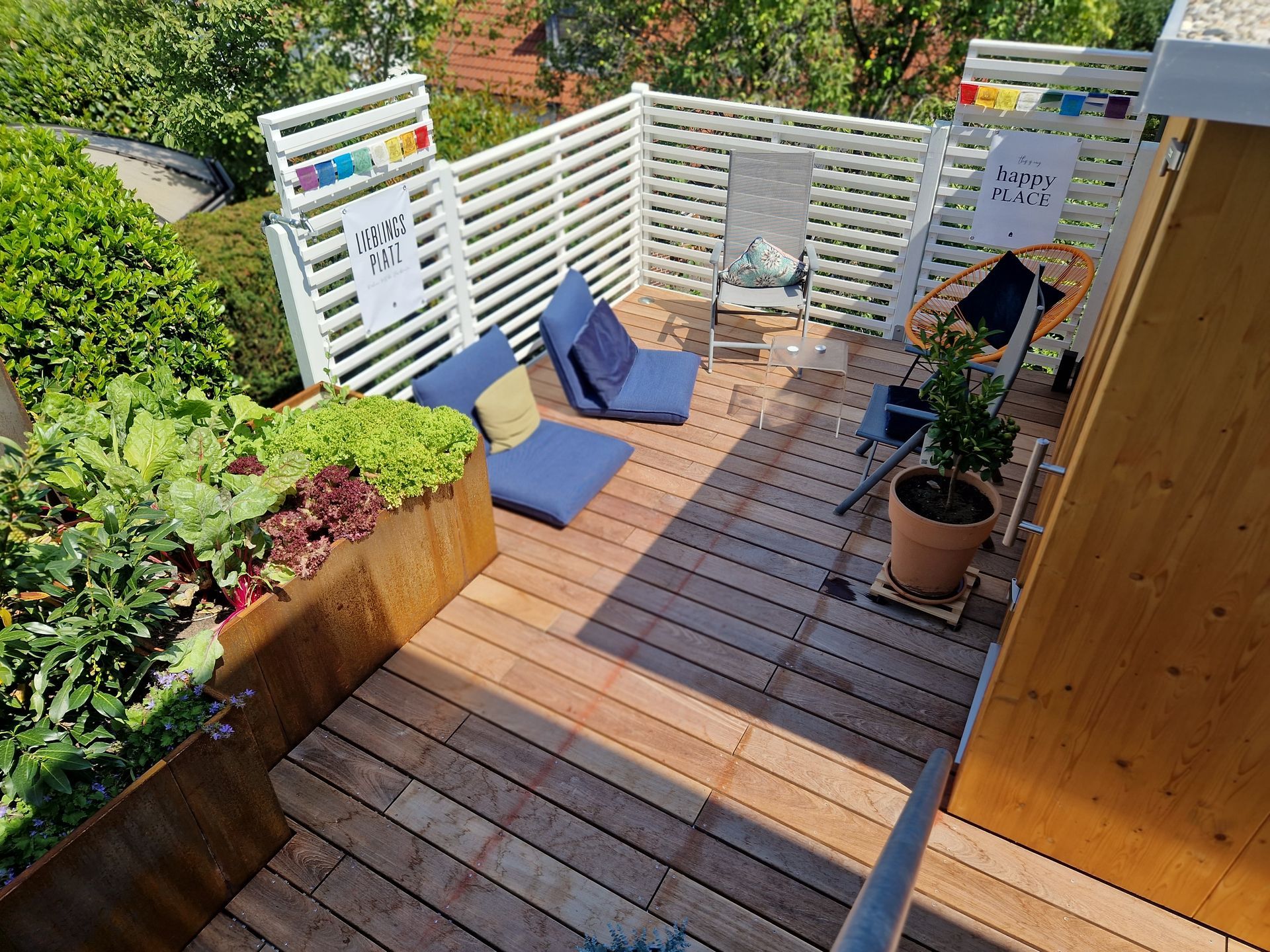 Holzterrasse mit blauen Kissen, weißer Lattenzaun und Kübelgarten.