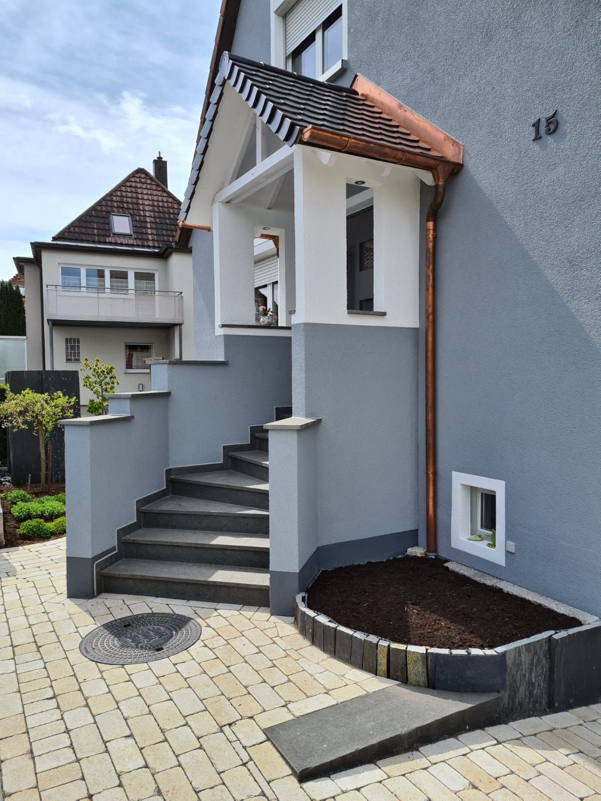 Eingang zu einem grauen Haus mit Treppe, Rampe und einer kleinen Veranda mit kupferverziertem Dach.