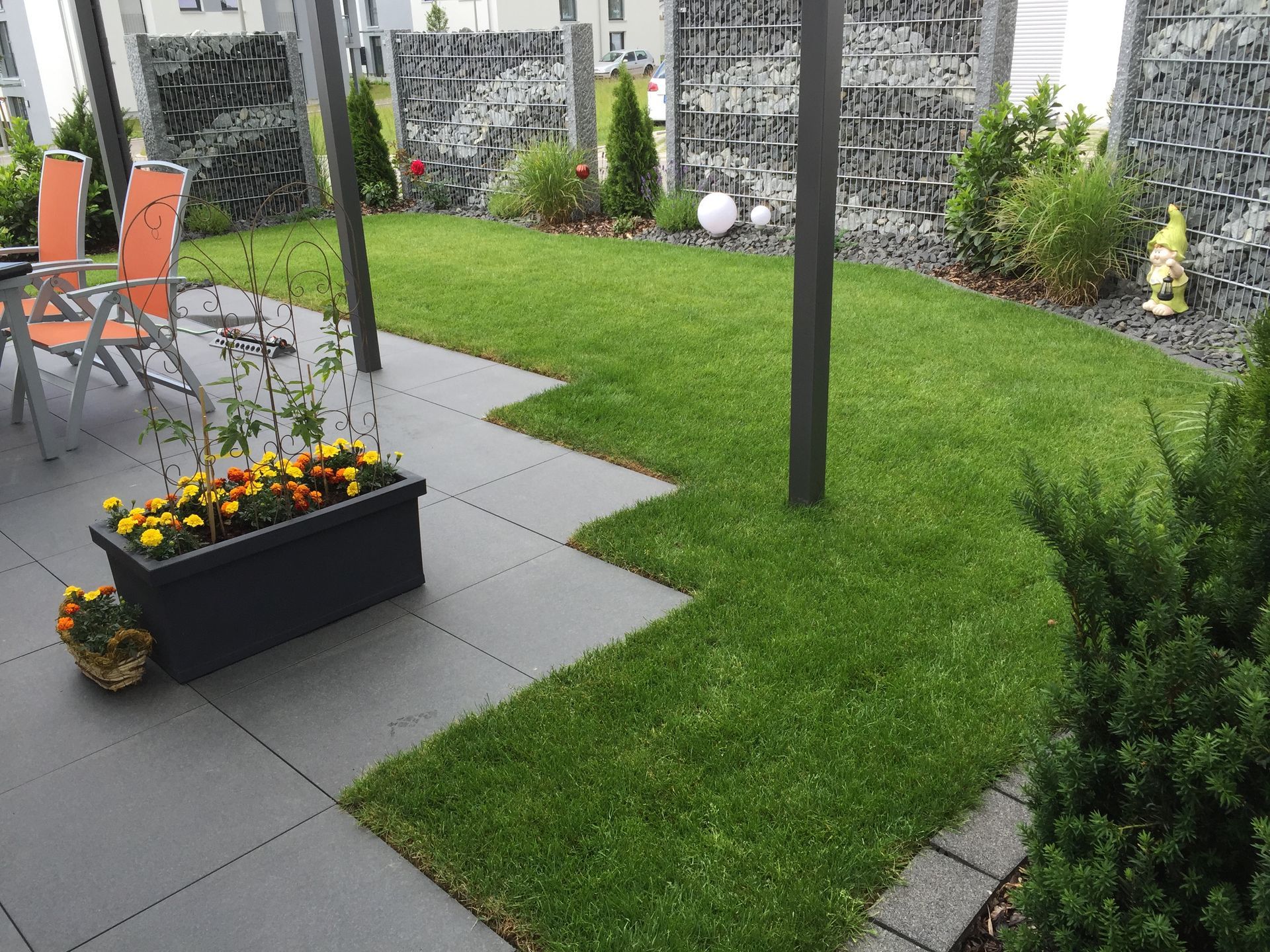 Terrasse mit grauen Fliesen, grünem Rasen, einem Gartenbeet mit gelben Blumen und einer Gabionenmauer.