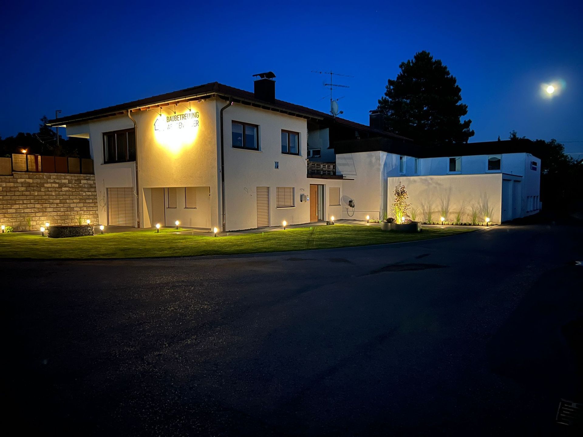 Ein zweistöckiges weißes Haus bei Nacht, beleuchtet von Bodenleuchten. Ein helles Schild ist im Obergeschoss zu sehen.