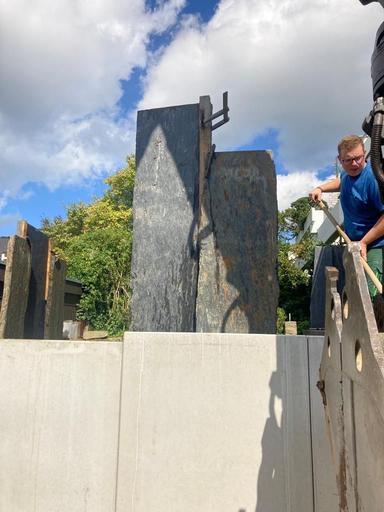 Ein Mann weist einen Kran ein, der eine große, dunkle Steinskulptur in einem von Bäumen umgebenen Hof unter bewölktem Himmel anhebt.