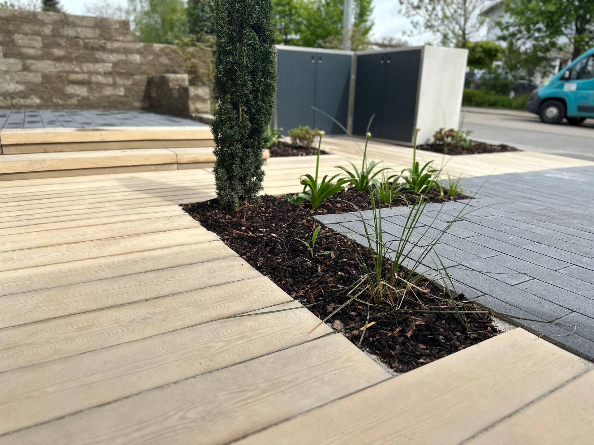 Ein Steinweg führt zu einem grasbewachsenen Hof mit einem Baum und Sträuchern, der von einer Holzterrasse aus zu sehen ist.