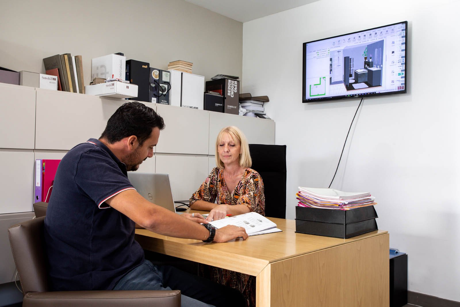 Un homme et une femme à leur bureau, consultant des documents. Un écran mural affiche un dessin. Environnement de bureau.