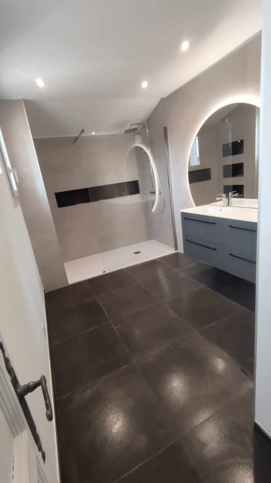 Salle de bain moderne avec carrelage gris, douche à l'italienne, sol foncé, miroir rond éclairé et vanité grise.