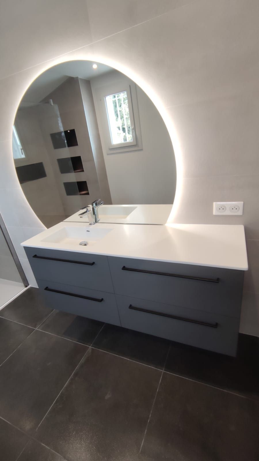 Salle de bain moderne avec une vanité grise, un grand miroir rond avec lumières et du carrelage noir.