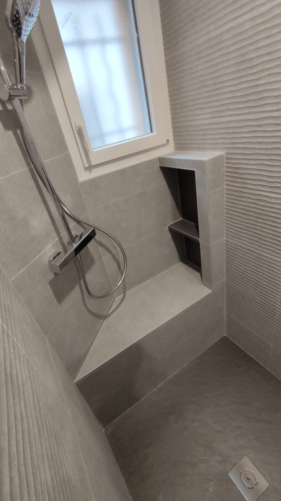 Douche carrelée grise avec banc intégré, étagères en alcôve et fenêtre. Pommeau et bonde de douche apparents.