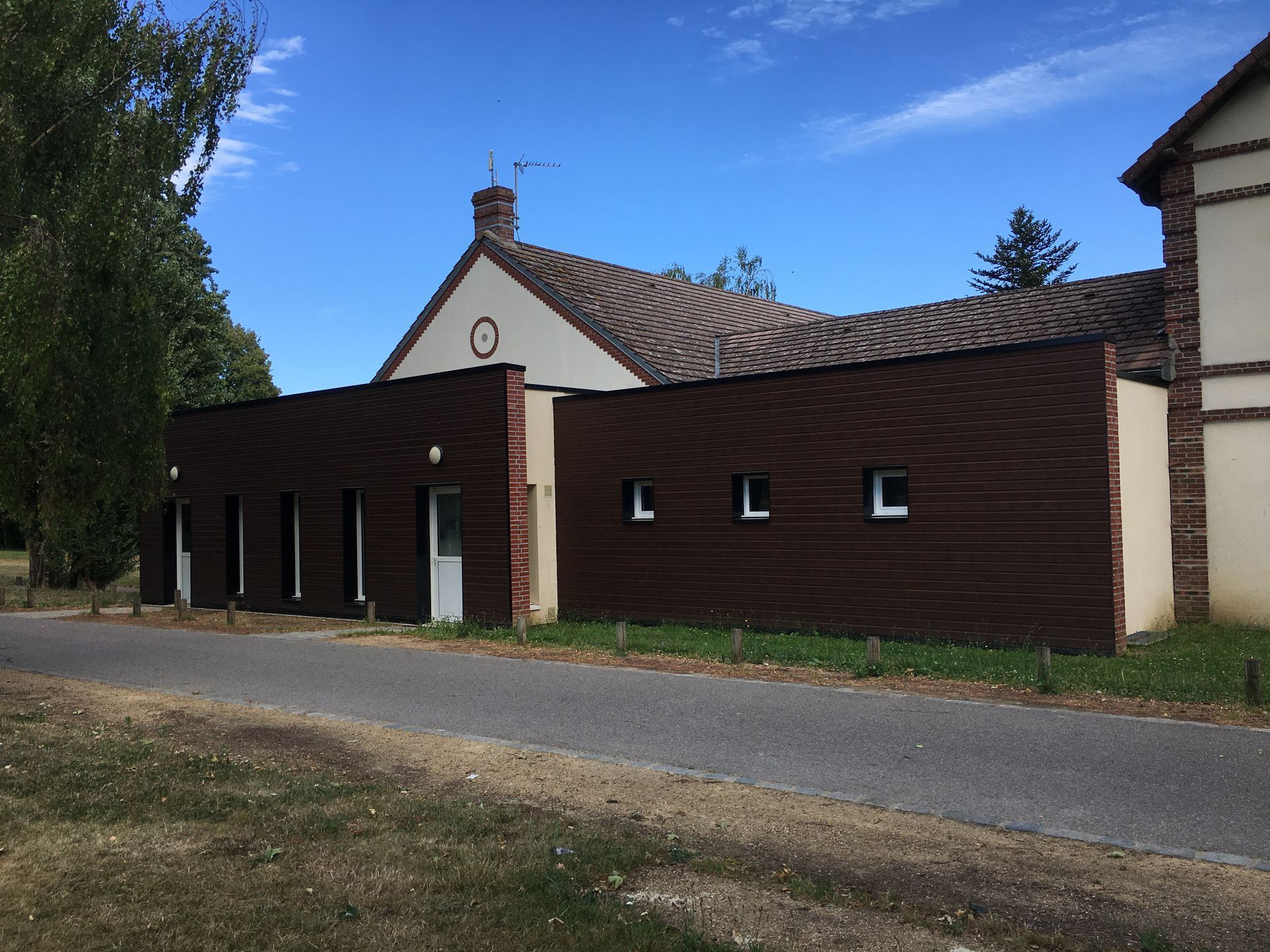 Un bâtiment beige avec une extension extérieure à lattes horizontales sombres, un toit à pignon et une route au premier plan.