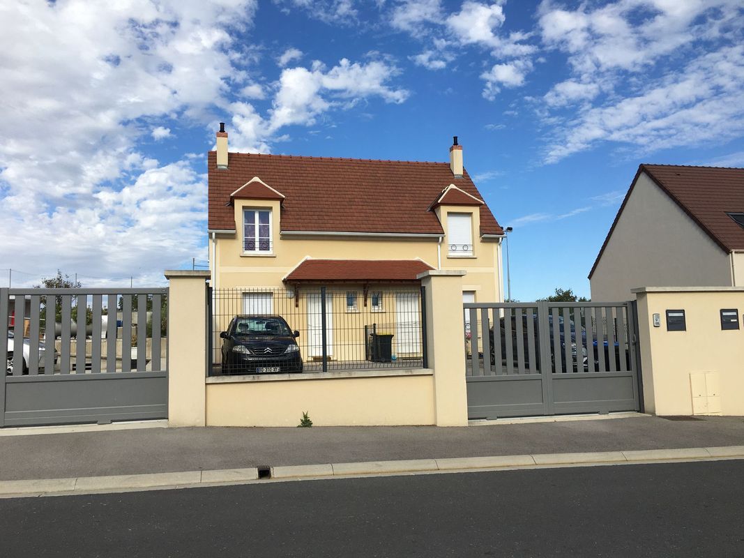 Une maison jaune clair à deux étages, avec un toit de tuiles brunes et des lucarnes, derrière une clôture et un portail gris.
