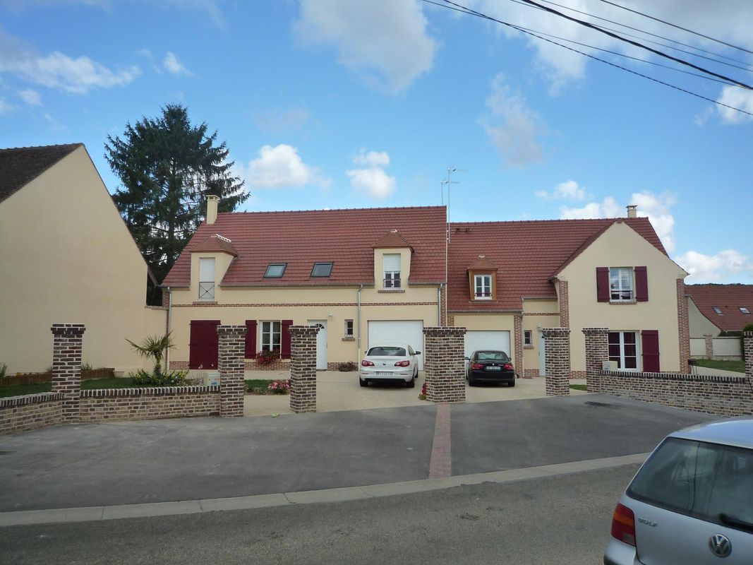 Un immeuble résidentiel beige de deux étages avec un toit de tuiles rouges, une allée d'accès et deux voitures garées.