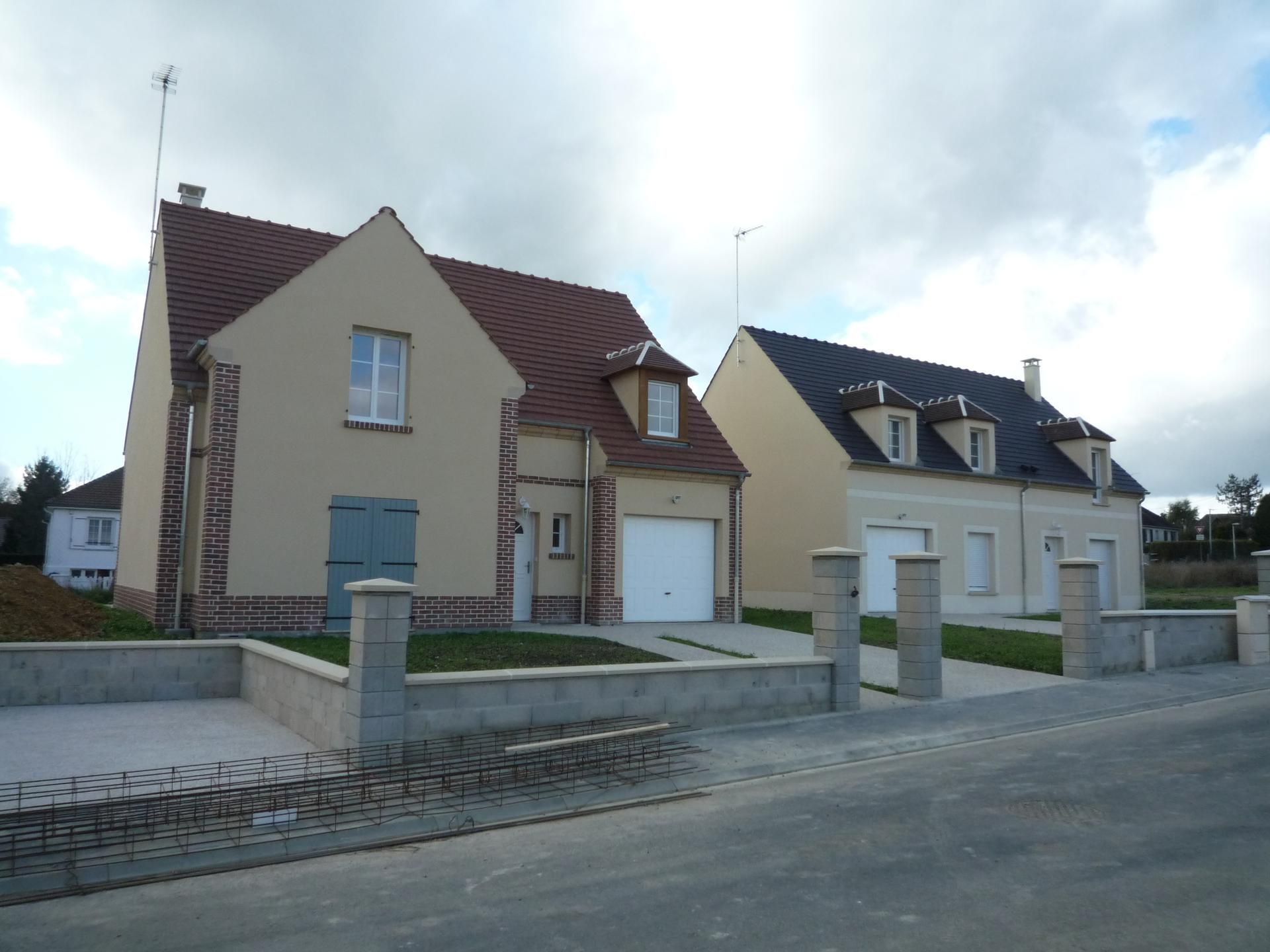 Deux maisons de banlieue nouvellement construites, aux murs en stuc beige et aux toits de tuiles sombres, se trouvent derrière un muret en béton.