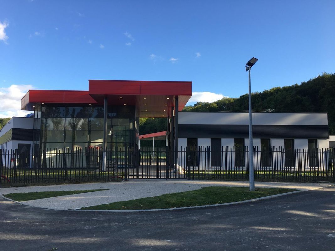 Un bâtiment moderne doté d'un auvent rouge, d'une façade en verre et de murs blancs, entouré d'une clôture métallique noire.