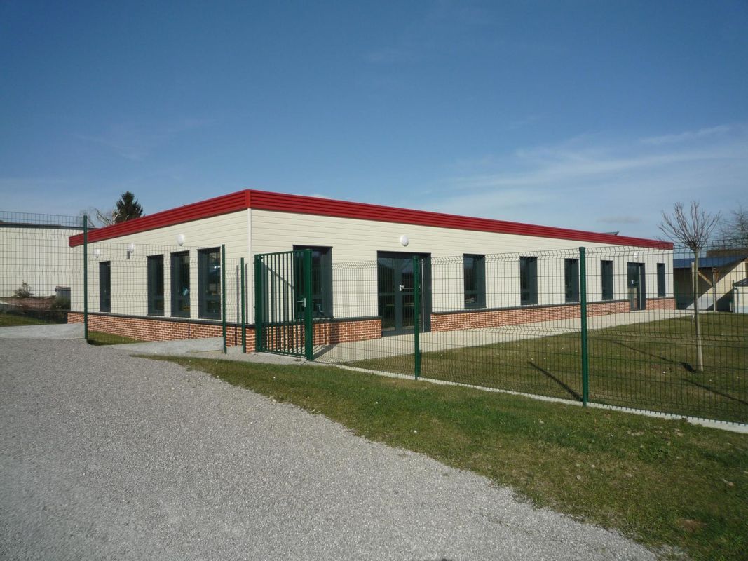 Un bâtiment rectangulaire moderne de plain-pied, aux murs clairs, avec une toiture rouge et des fenêtres sombres, situé sur un terrain gravillonné.
