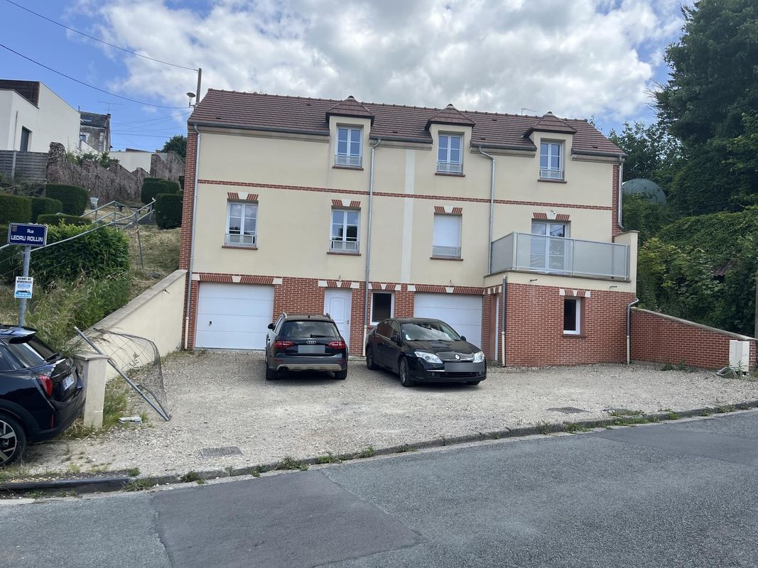 Une maison de ville beige de trois étages avec deux portes de garage blanches, un balcon et deux voitures garées devant.