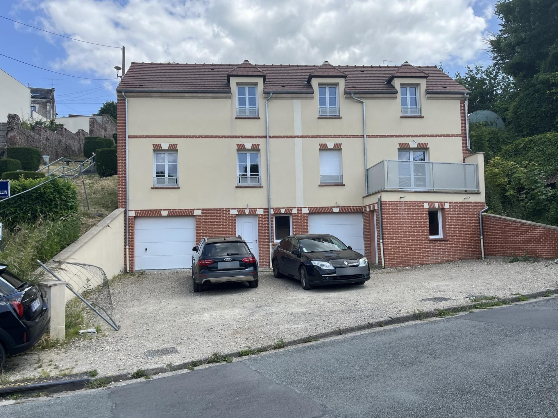 Un immeuble à plusieurs étages avec une façade claire, un toit brun, un rez-de-chaussée en briques et deux garages.