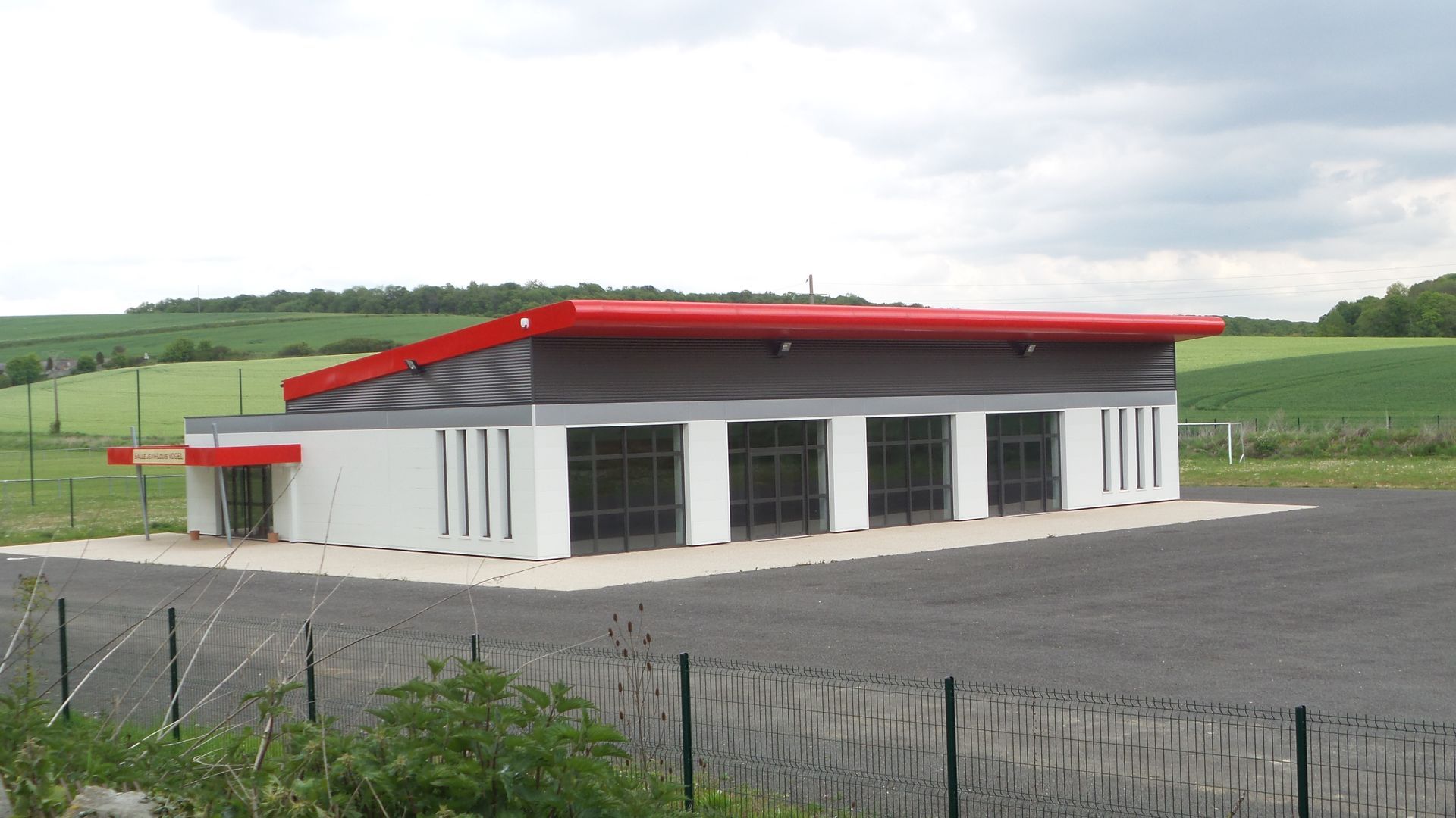Un bâtiment industriel moderne de plain-pied, avec un toit rouge, des boiseries sombres et des murs blancs.