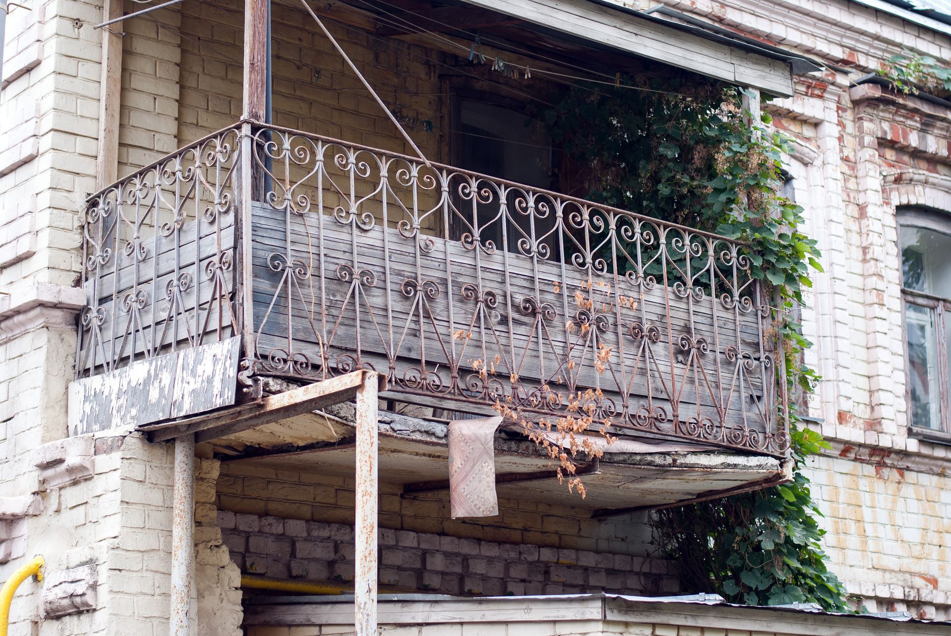 Un balcon en fer forgé, patiné par le temps et orné, est adossé à un bâtiment en briques claires délabré, sur lequel poussent des vignes.