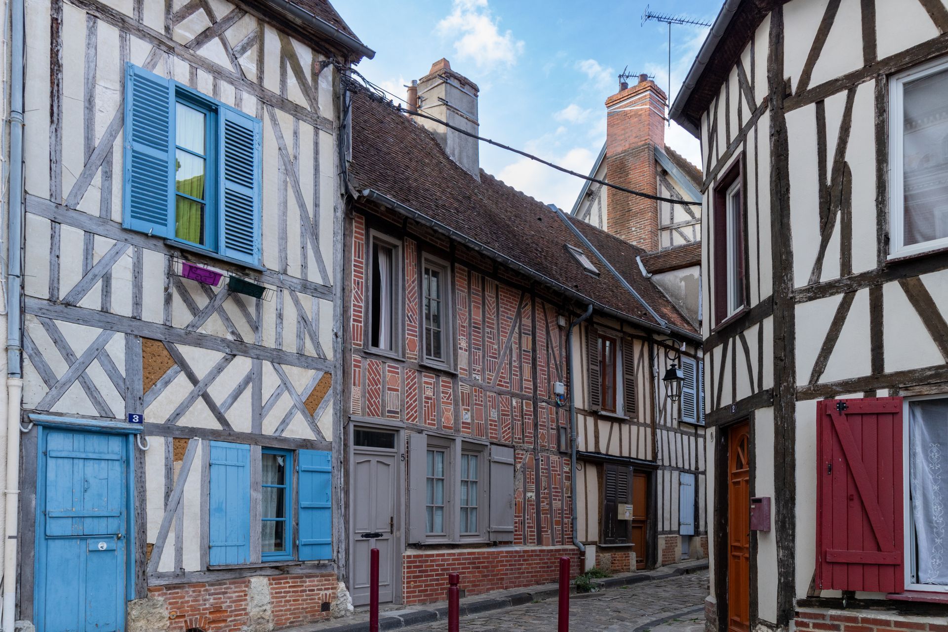 Une rue pavée étroite bordée de maisons historiques à colombages aux volets bleus, bruns et rouges.