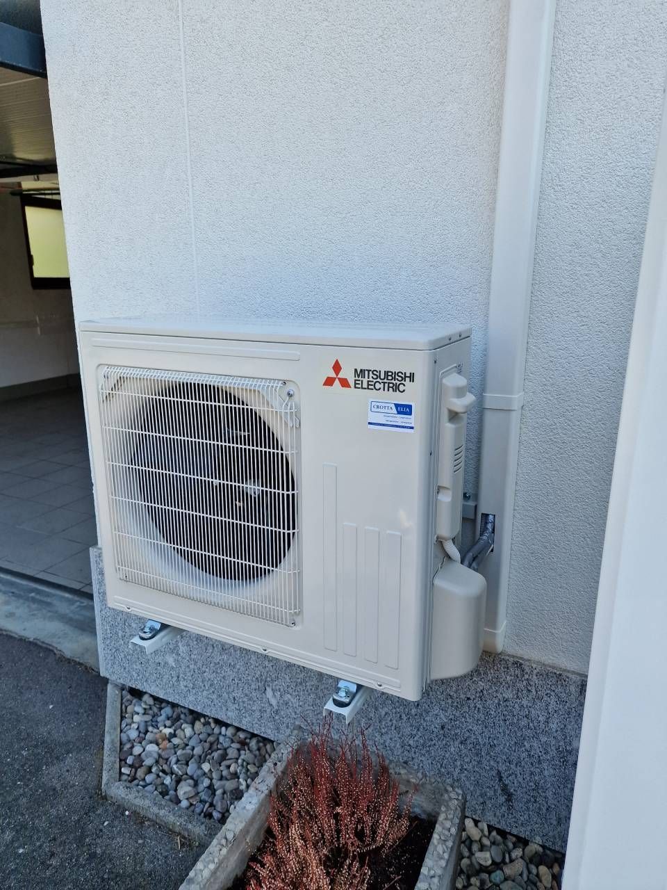 Unità esterna di condizionamento dell'aria Mitsubishi montata su una parete accanto a un letto di ghiaia e alla facciata esterna dell'edificio.