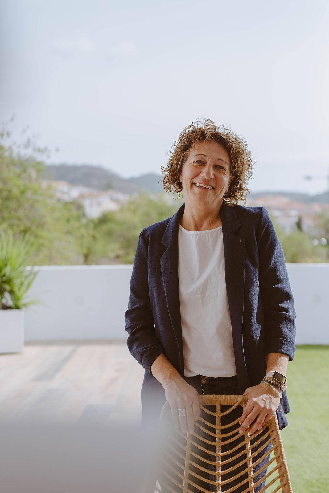 Una mujer con un blazer azul marino sonríe, parada cerca de una silla en un patio con vista a un pueblo con montañas de fondo