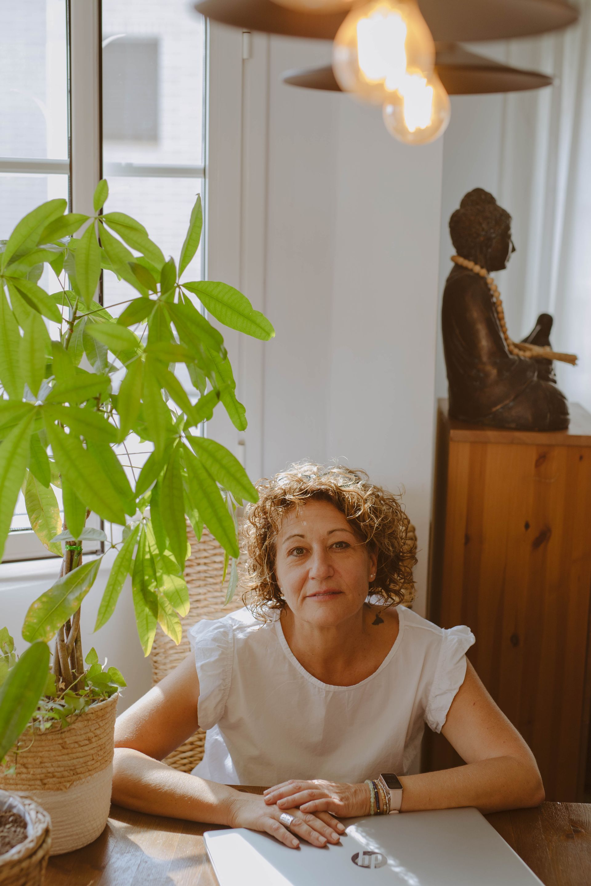 Una mujer de cabello rizado está sentada a una mesa, sonriendo. Detrás de ella, una planta, una estatua de Buda y una lámpara