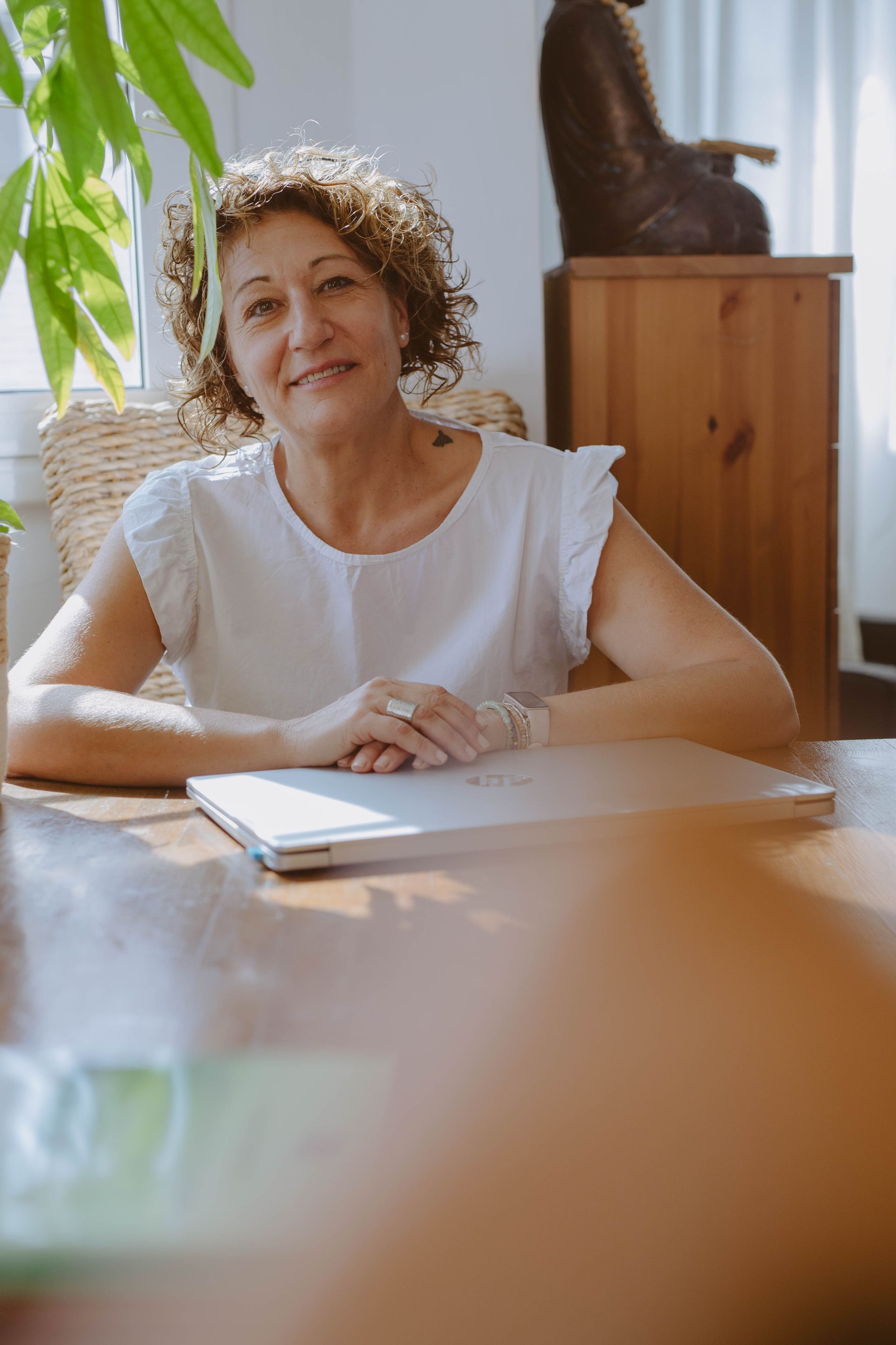 Mujer sentada en una mesa con un ordenador portátil, sonriendo, habitación soleada.