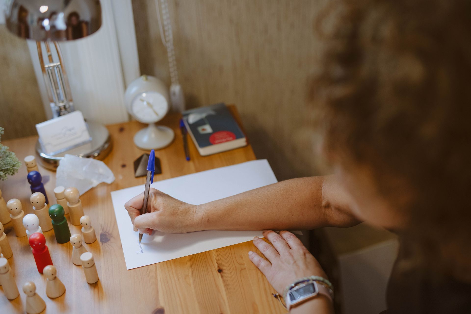 Persona escribiendo en papel en un escritorio de madera, con lámpara y reloj cerca.