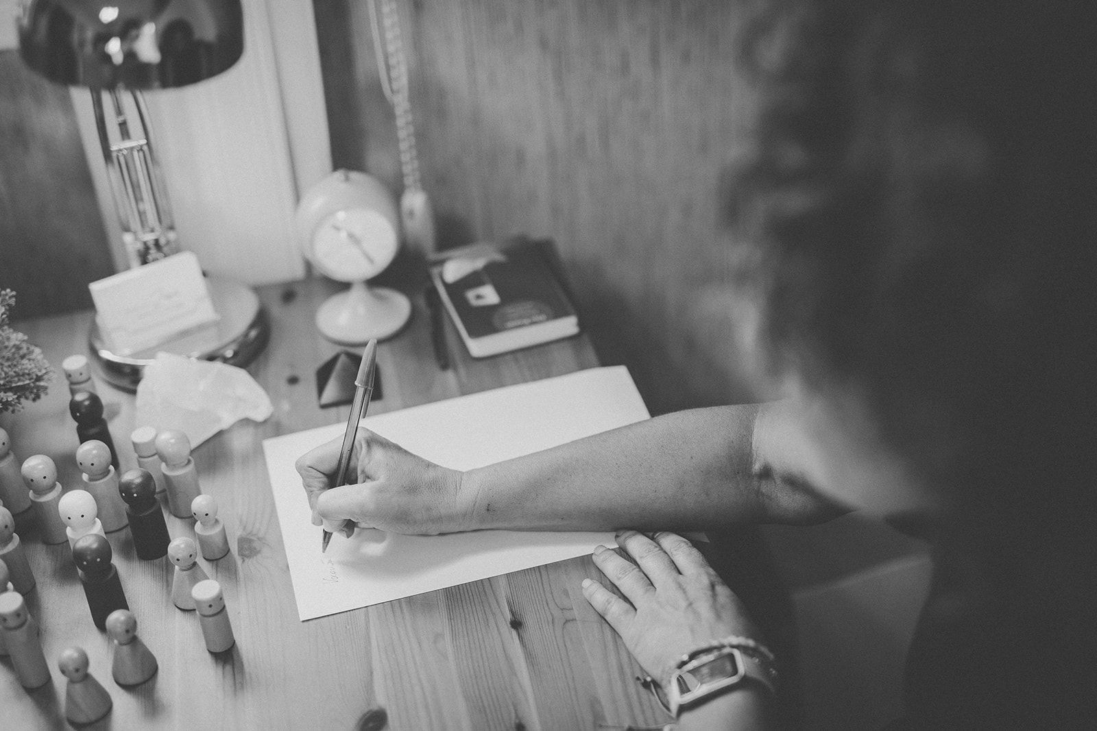 Una mujer con cabello rizado sonríe, apoyando la barbilla en la mano en una mesa, mirando hacia otra persona.