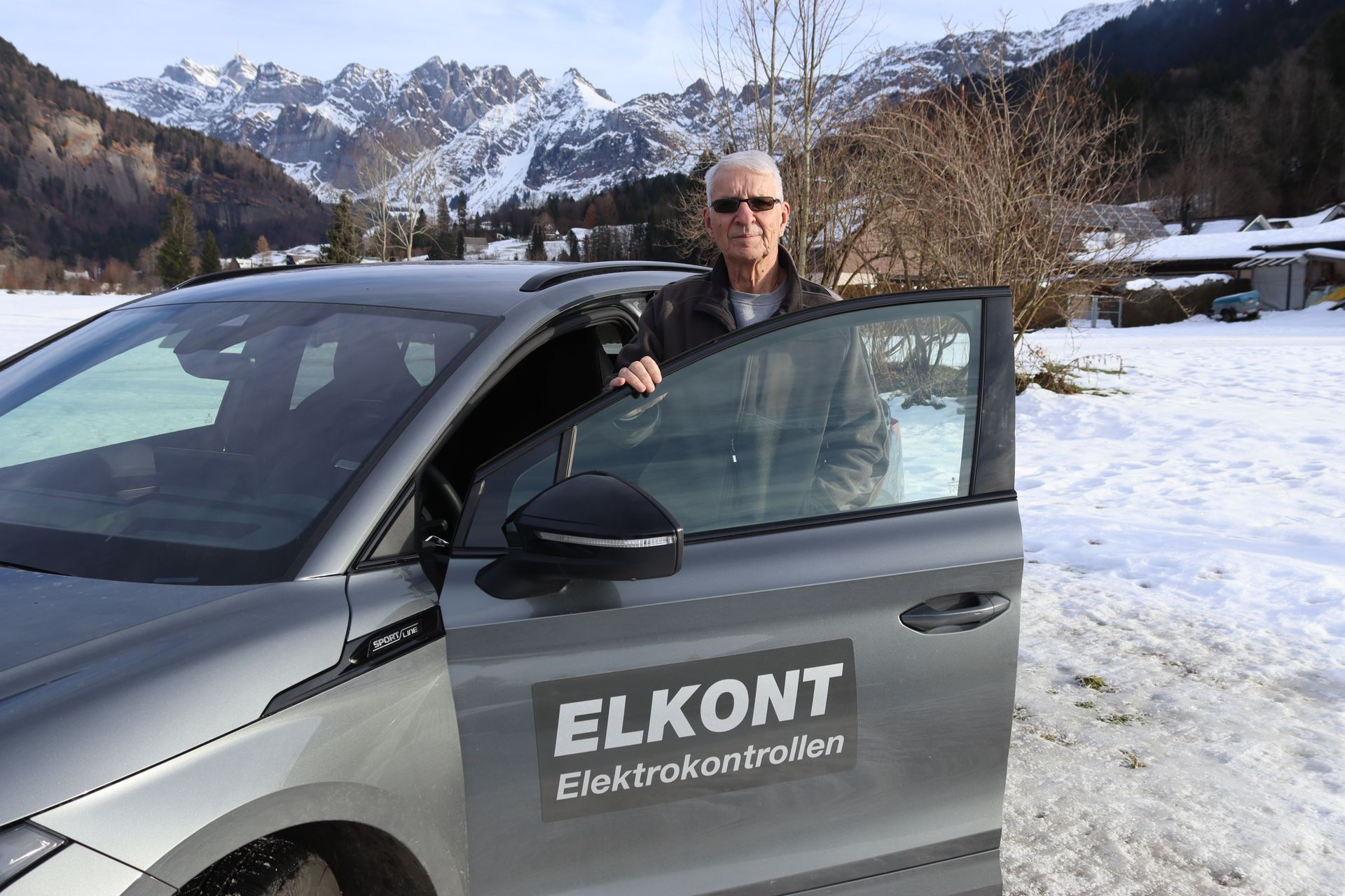 Ein Mann steht neben einem grauen Auto mit dem Logo „ELKONT“, im Hintergrund sind Berge zu sehen, auf dem Boden liegt Schnee.