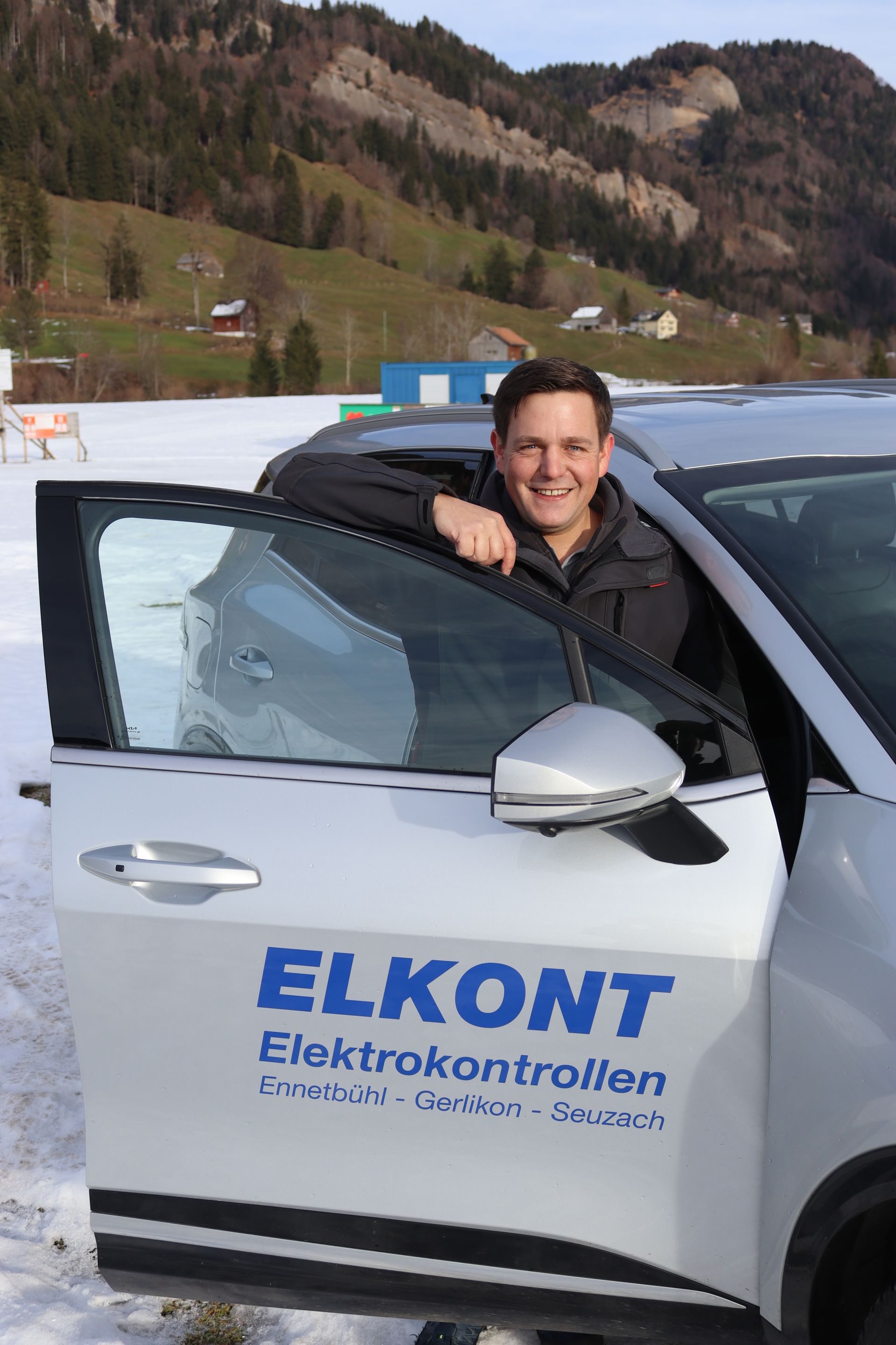 Mann in einem Auto mit dem Logo „ELKONT Elektrokontrollen“, verschneite Landschaft, Berge im Hintergrund.