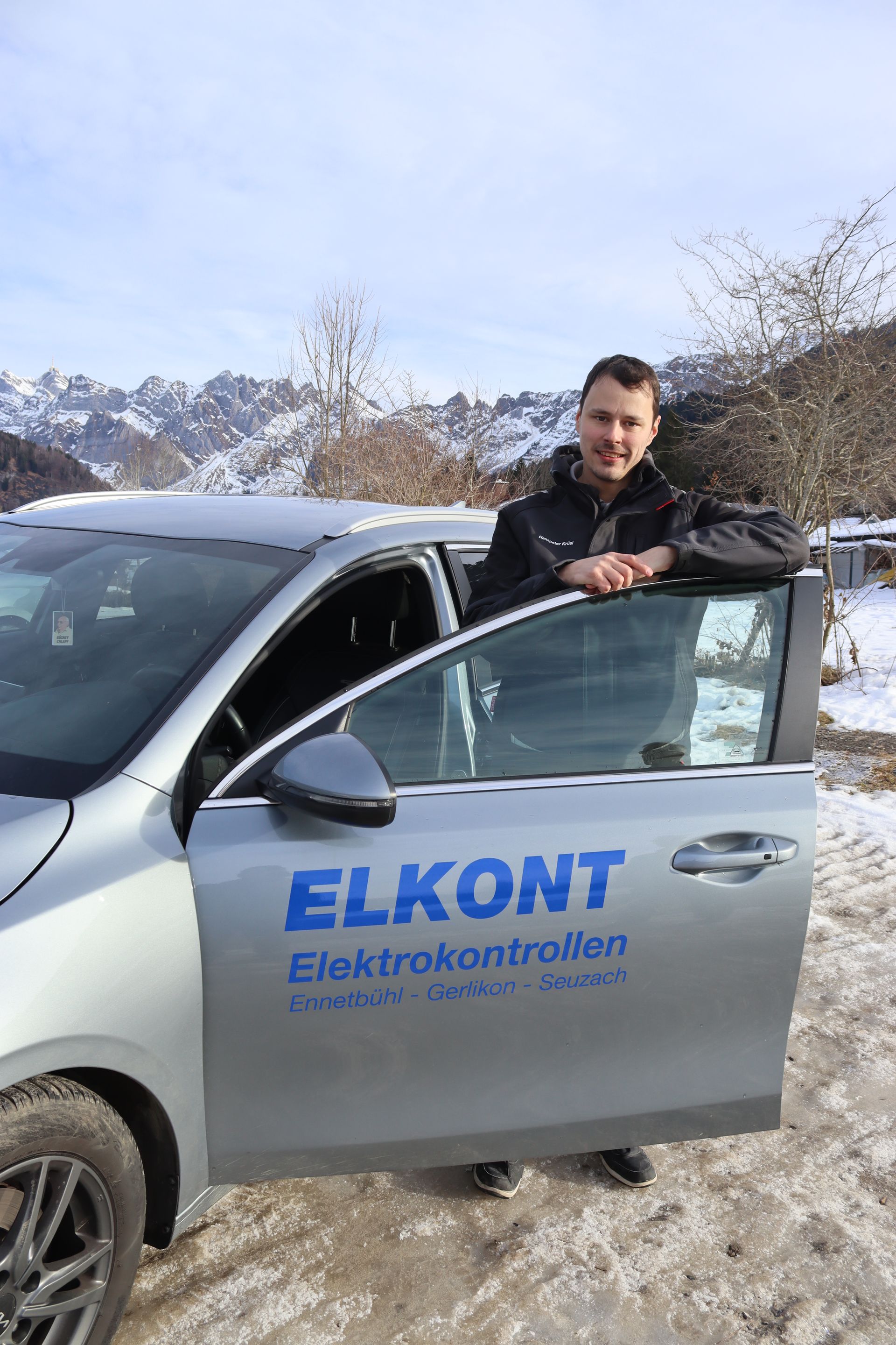 Mann im Auto mit dem Logo „ELKONT Elektrokonrtollen“, im Hintergrund schneebedeckte Berge.
