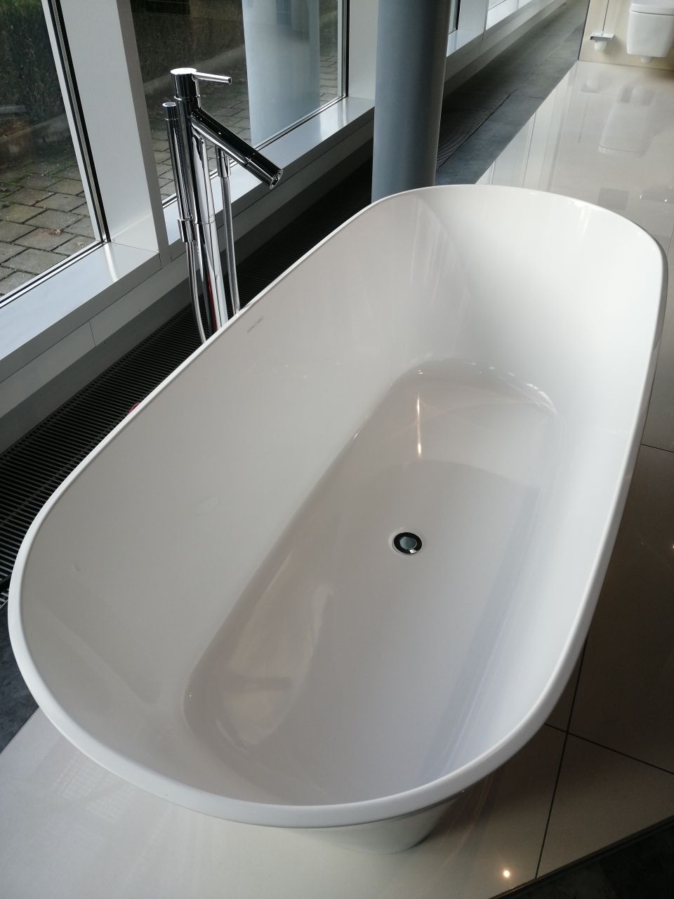 Eine weiße Badewanne steht auf einem gefliesten Boden neben einem Fenster.