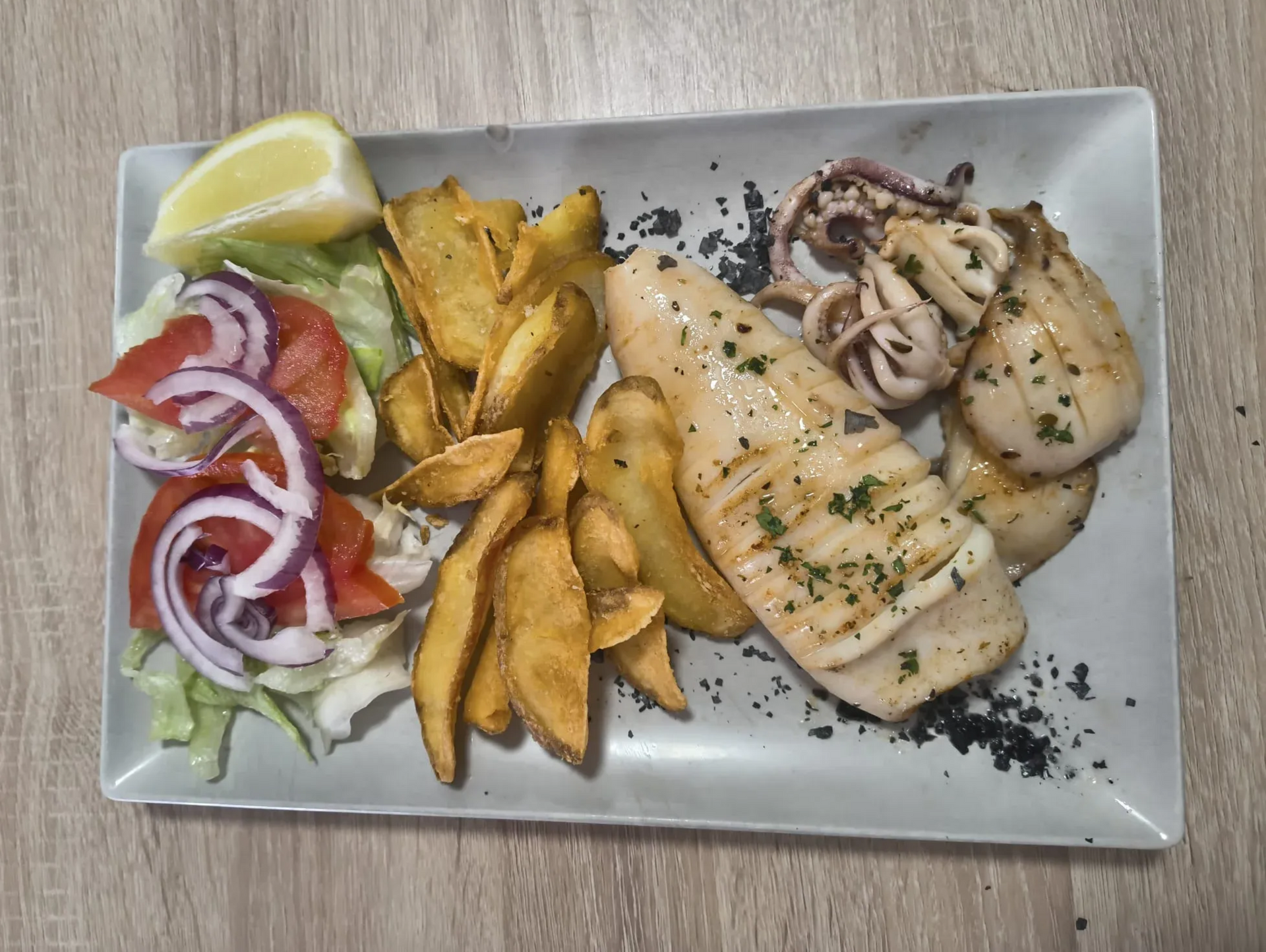 Pescado a la parrilla, calamares, patatas gajo y ensalada con limón en un plato rectangular.