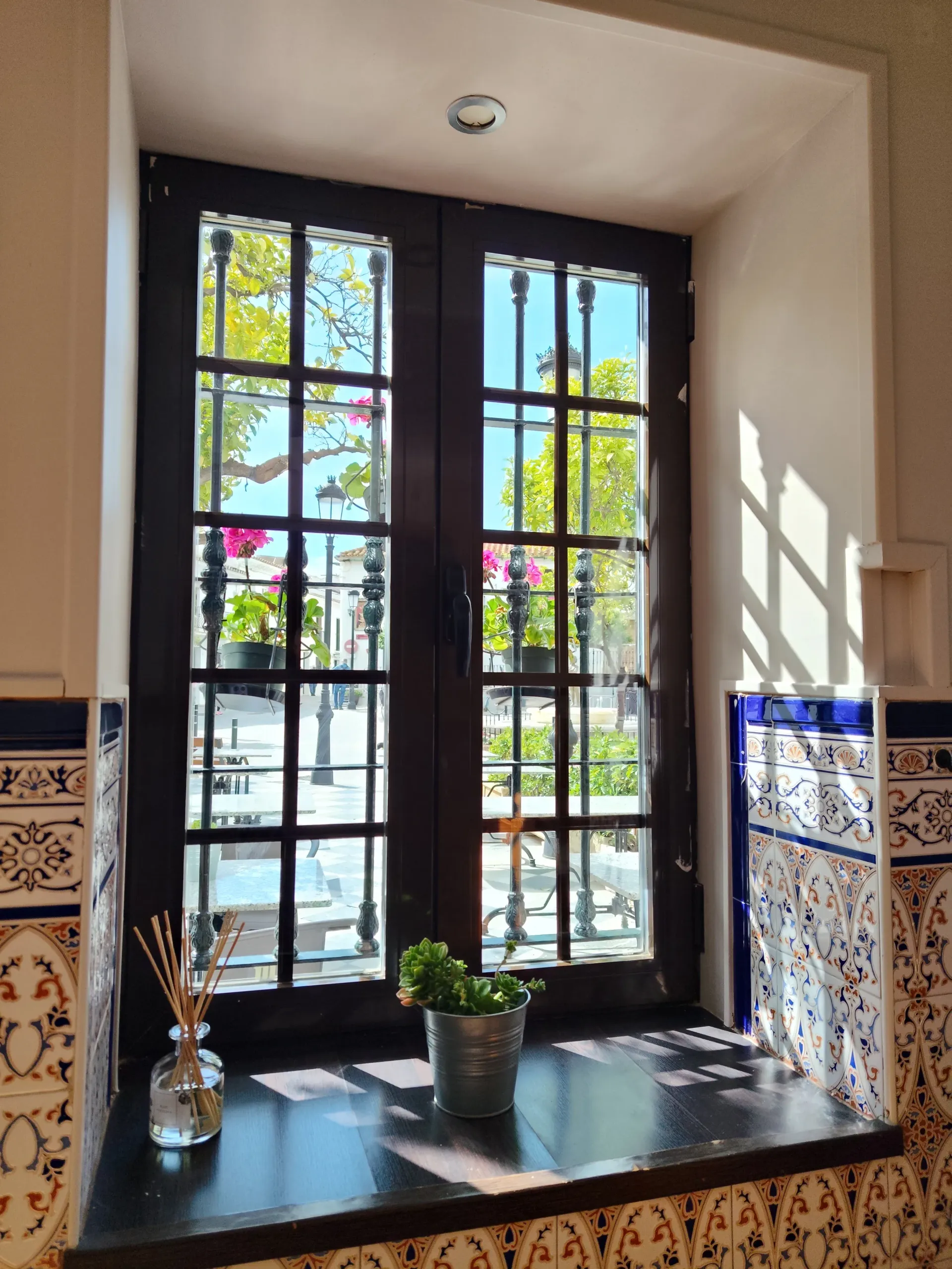Rincón soleado junto a la ventana con cristales enmarcados en negro, una planta en maceta y paredes de azulejos estampados.