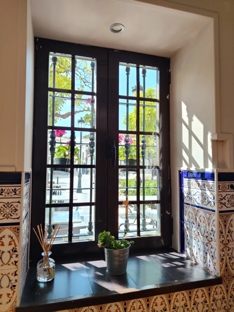 Rincón soleado junto a la ventana con cristales enmarcados en negro, una planta en maceta y paredes de azulejos estampados.