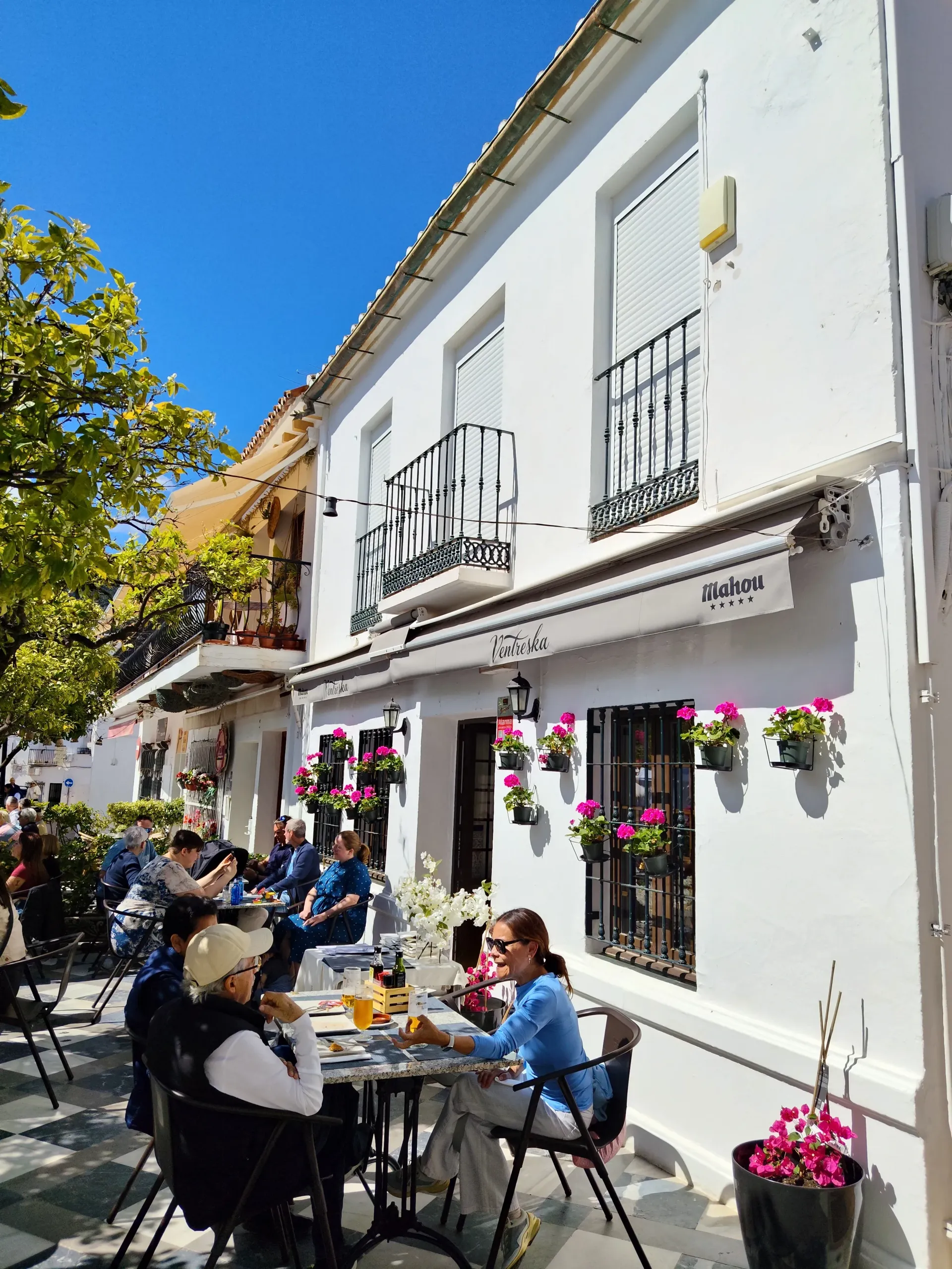 Cafetería al aire libre en una soleada calle blanca, con gente sentada en mesas y cestas de flores rosas colgando de la pared.