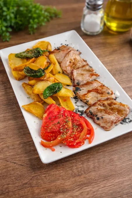 Carne a la parrilla con patatas asadas y tomates en un plato blanco, adornada con hierbas aromáticas.