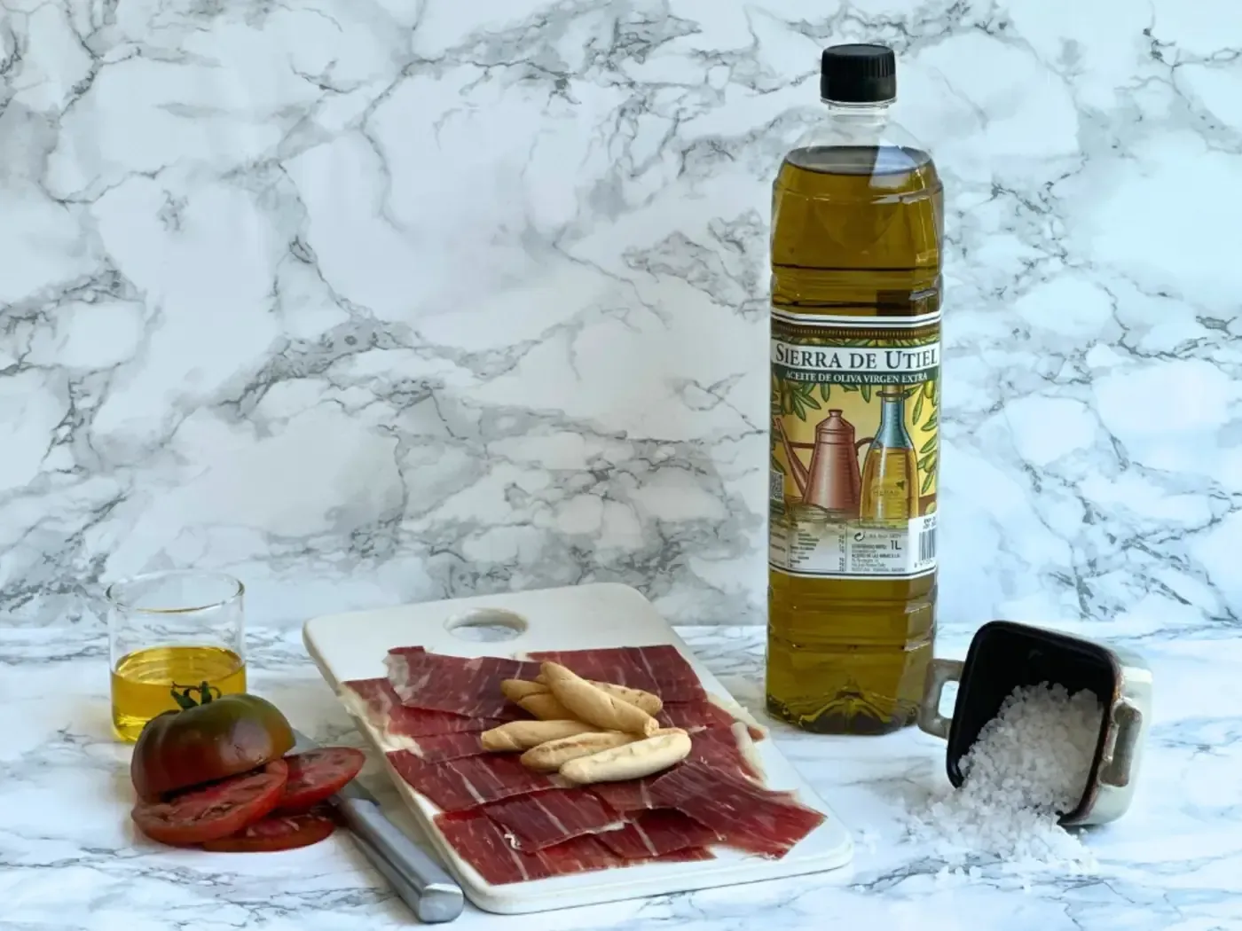 Jamón loncheado, tomates, aceite de oliva, palitos de pan, sal y botella de aceite de oliva sobre mármol.