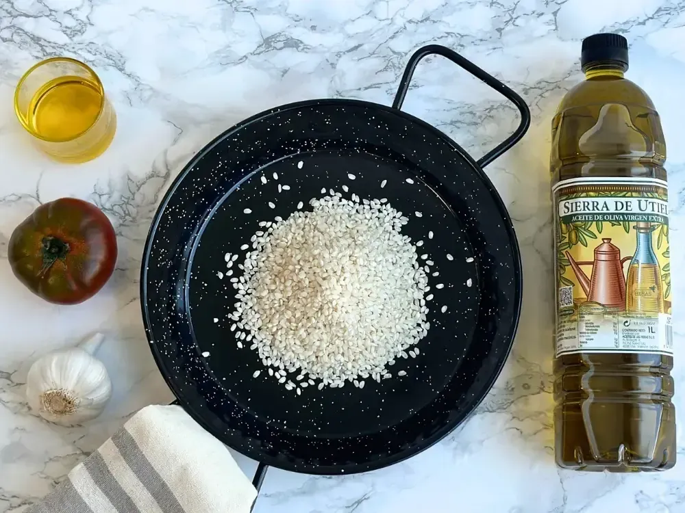 Ingredientes para la paella: arroz en una sartén, aceite de oliva, tomate, ajo y una botella de aceite sobre una superficie de mármol.