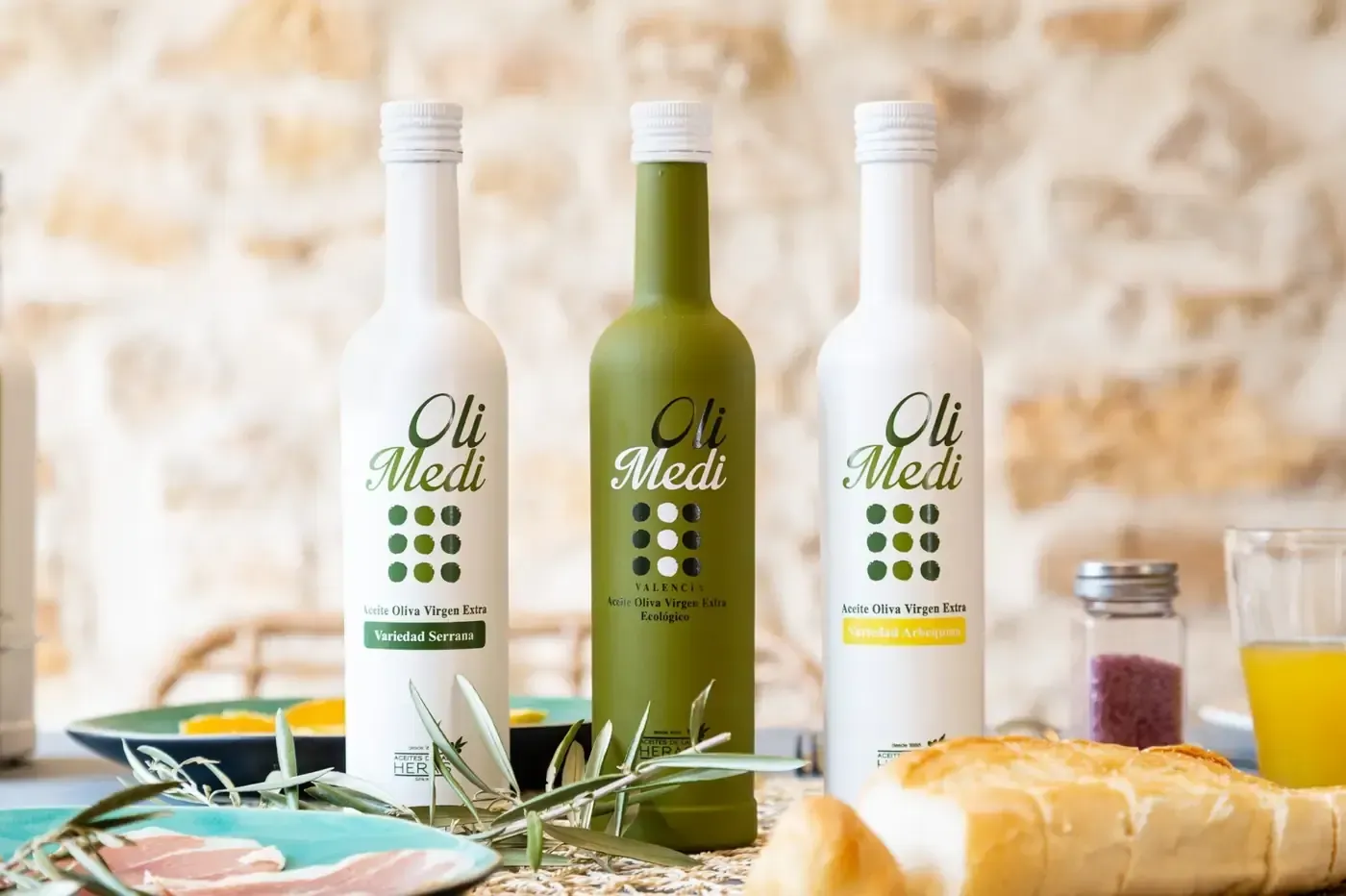 Tres botellas de aceite de oliva (Oli Medi) sobre una mesa con comida y un muro de piedra de fondo.