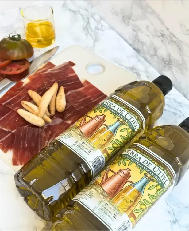 Botellas de aceite de oliva, embutidos, palitos de pan, tomates y un vasito de aceite sobre una superficie de mármol.