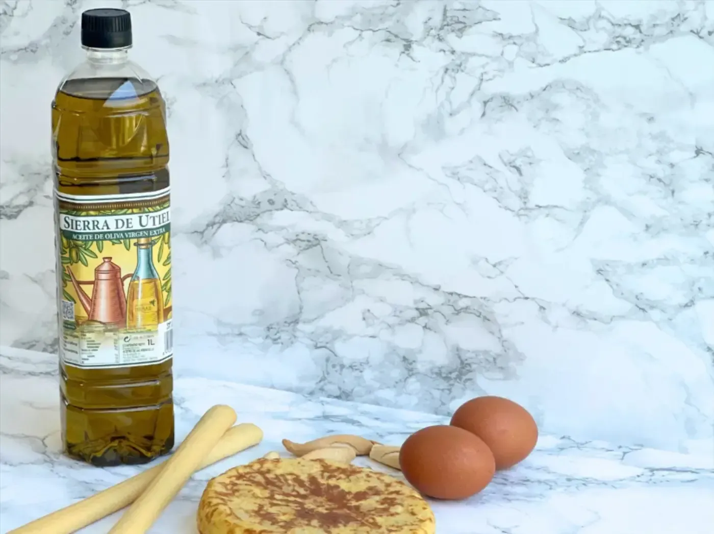Botella de aceite de oliva, dos huevos, tortilla de patatas y palitos de pan sobre una superficie de mármol.