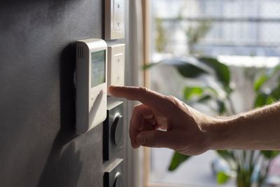 Une main actionne un thermostat mural dans une pièce ensoleillée, avec une plante et une fenêtre en arrière-plan.
