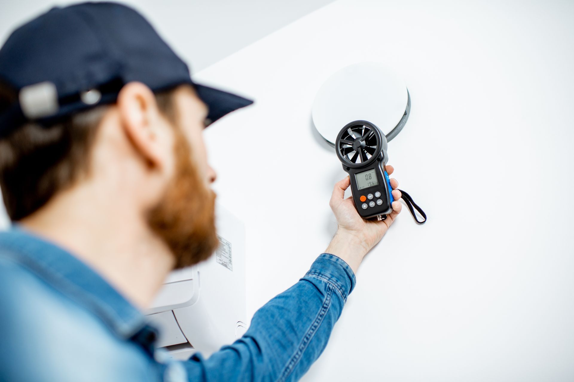 Un technicien inspecte un thermostat mural à l'aide d'un appareil de mesure portatif dans une pièce blanche.