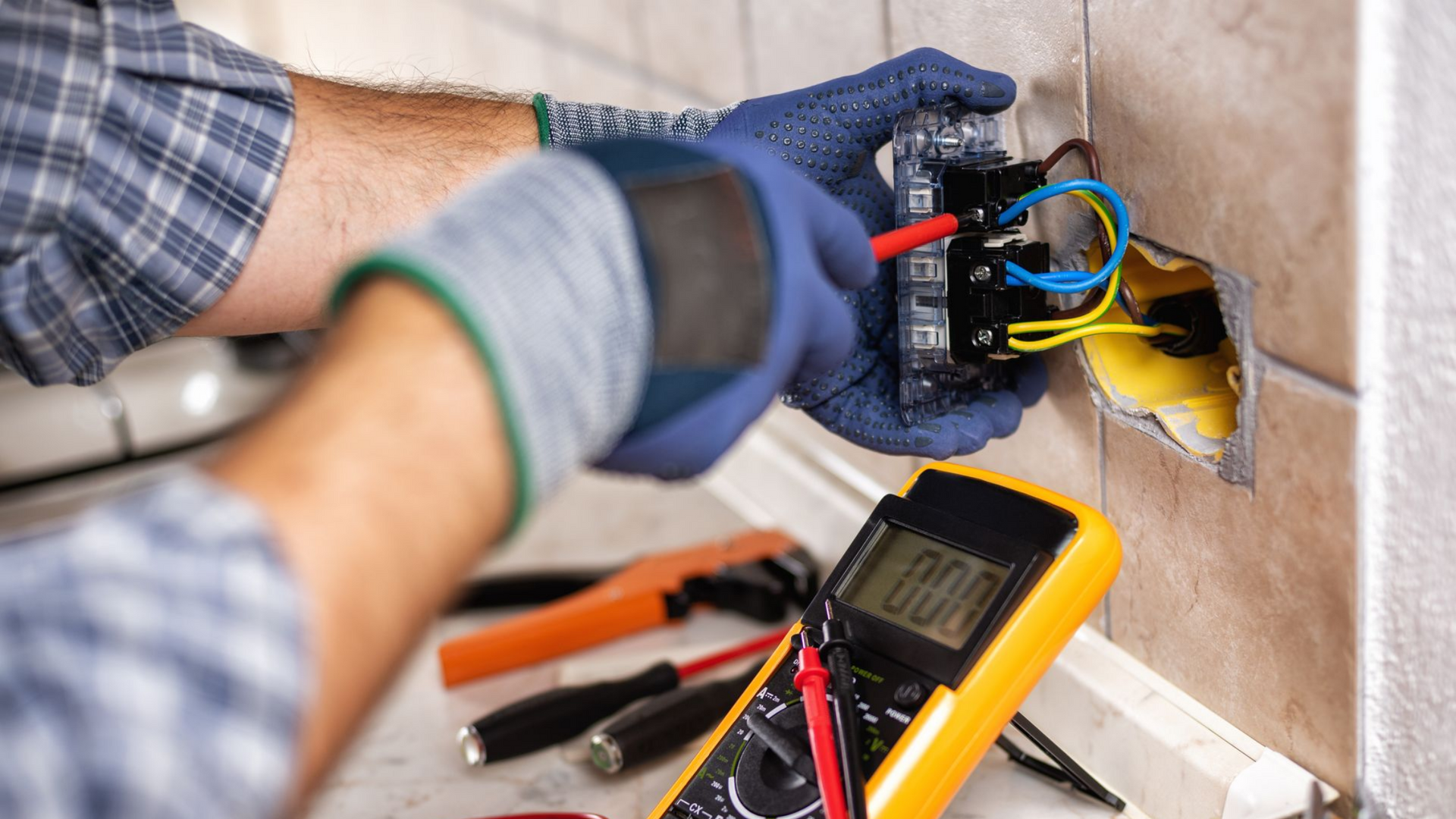 Un électricien teste les fils électriques apparents d'un mur à l'aide d'un multimètre et d'outils à proximité.