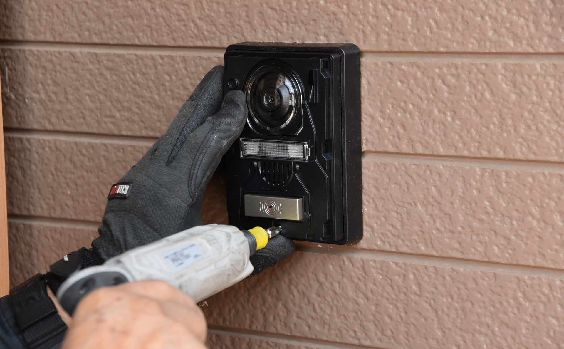 Main gantée utilisant un tournevis pour installer une caméra de sécurité extérieure noire sur un revêtement marron