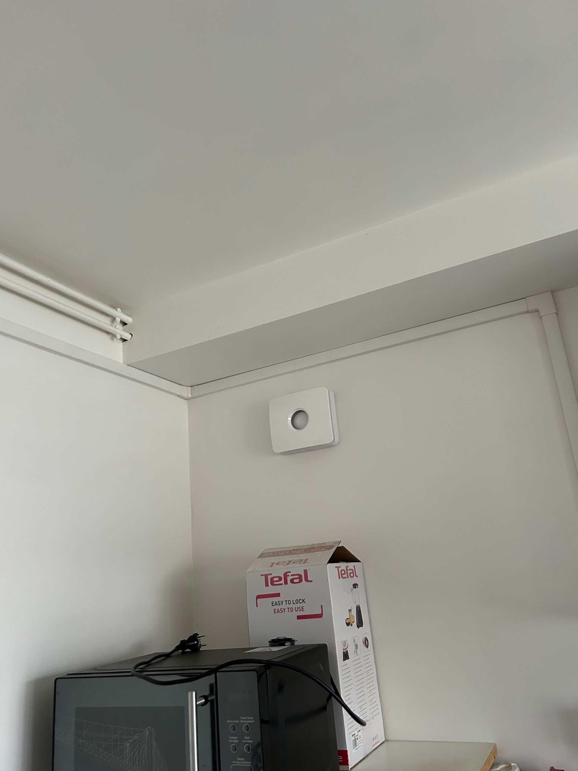 Coin d'une pièce avec une grille d'aération blanche au-dessus d'une armoire noire et d'une boîte en carton dessus.