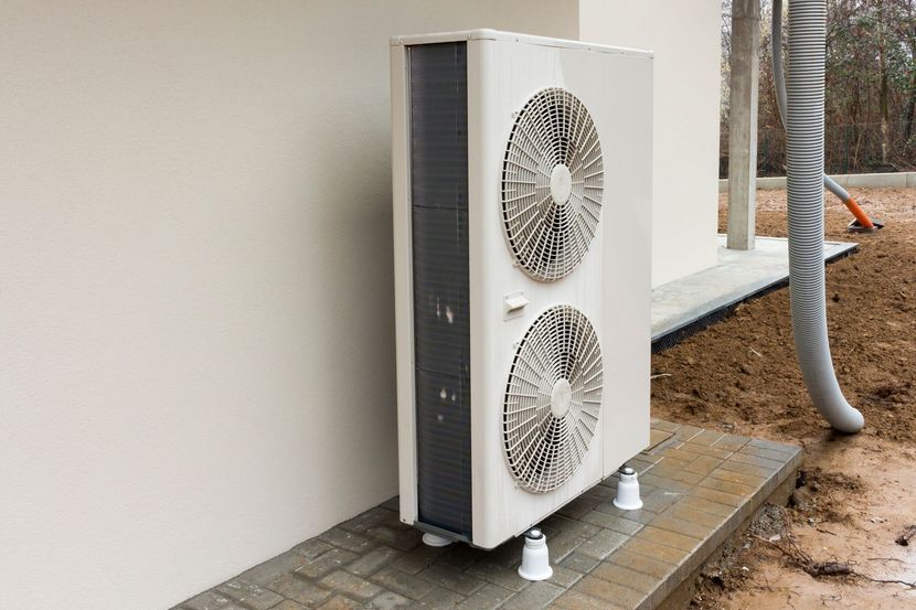 Unité de climatisation extérieure avec deux ventilateurs, fixée contre un mur sur une dalle de béton avec tuyaux apparents.