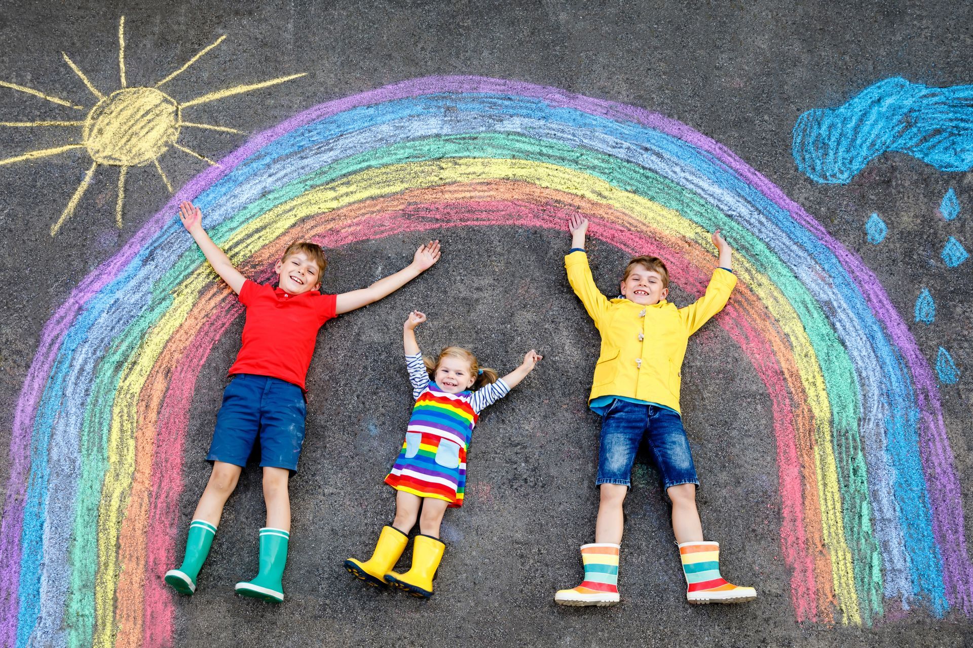 Trois enfants allongés dans une cour d'école sous un arc-en-ciel tracé à la craie