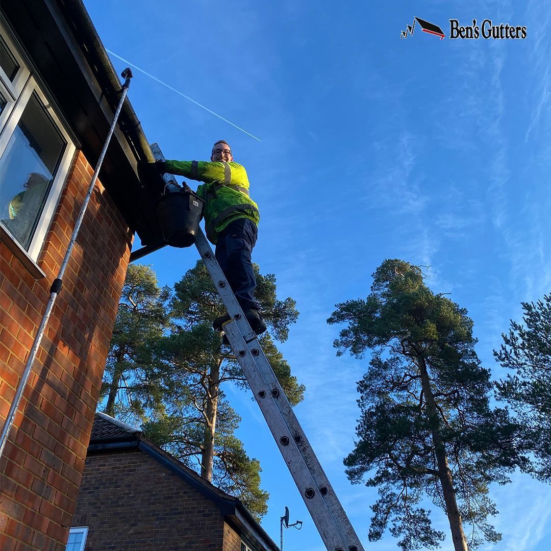 Worker on a ladder cleaning the upper side of a brick house against a clear blue sky.