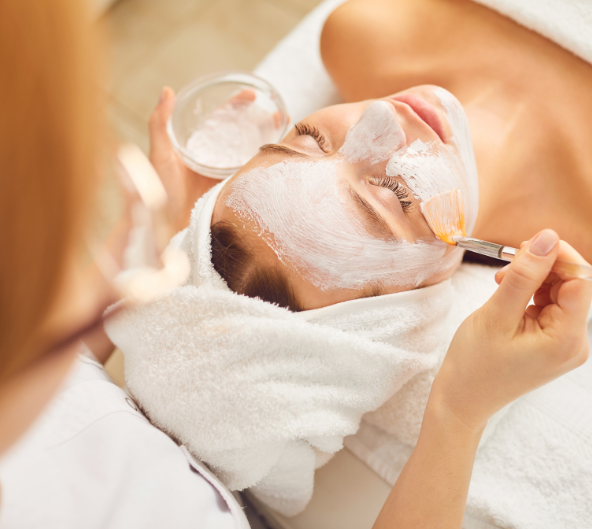 Une femme reçoit un masque facial dans un spa ; une esthéticienne applique le masque sur le visage avec un pinceau.